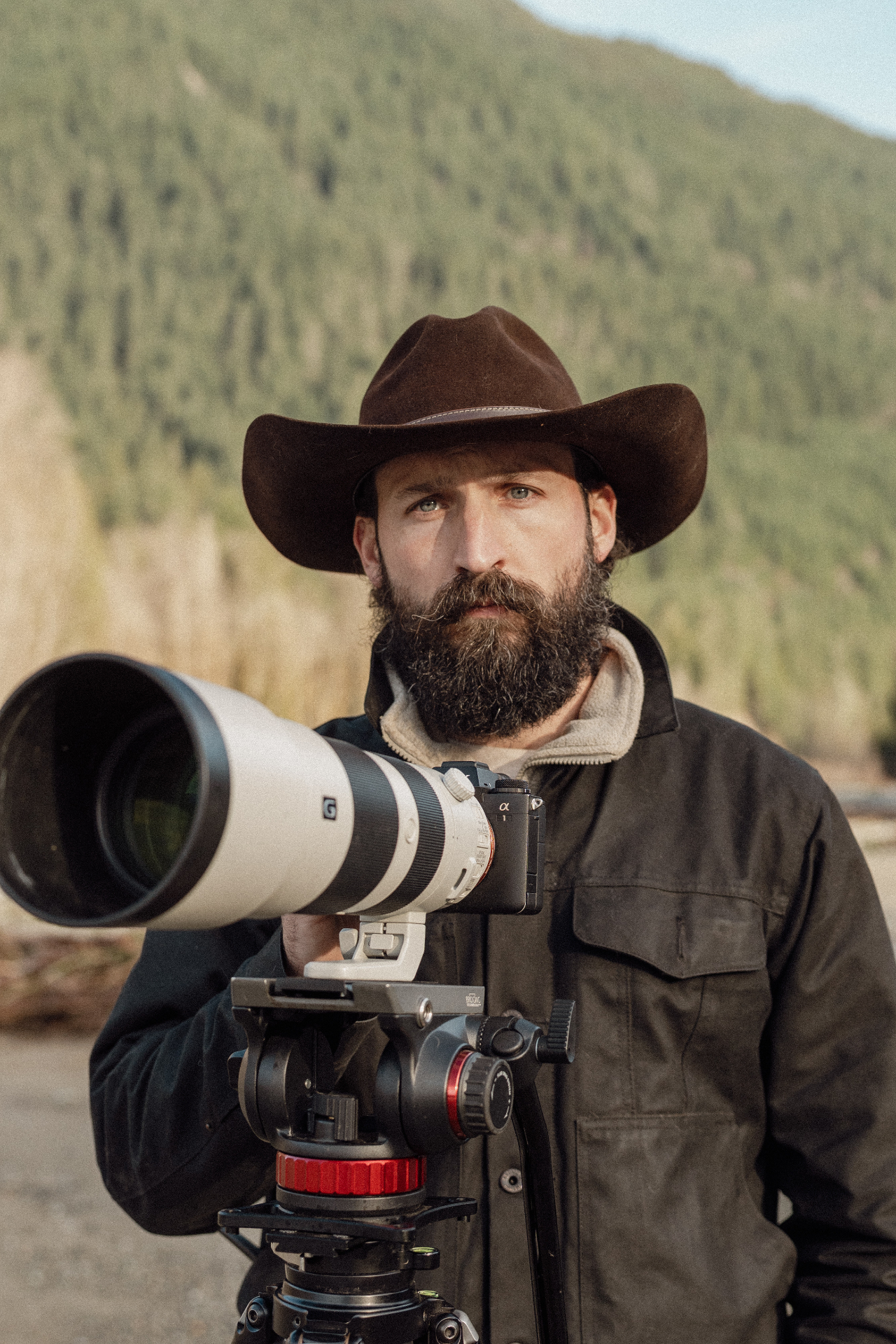 Portrait of Devin Groody in a wide-brim hat, standing behind a long telephoto lens with forested mountains behind him