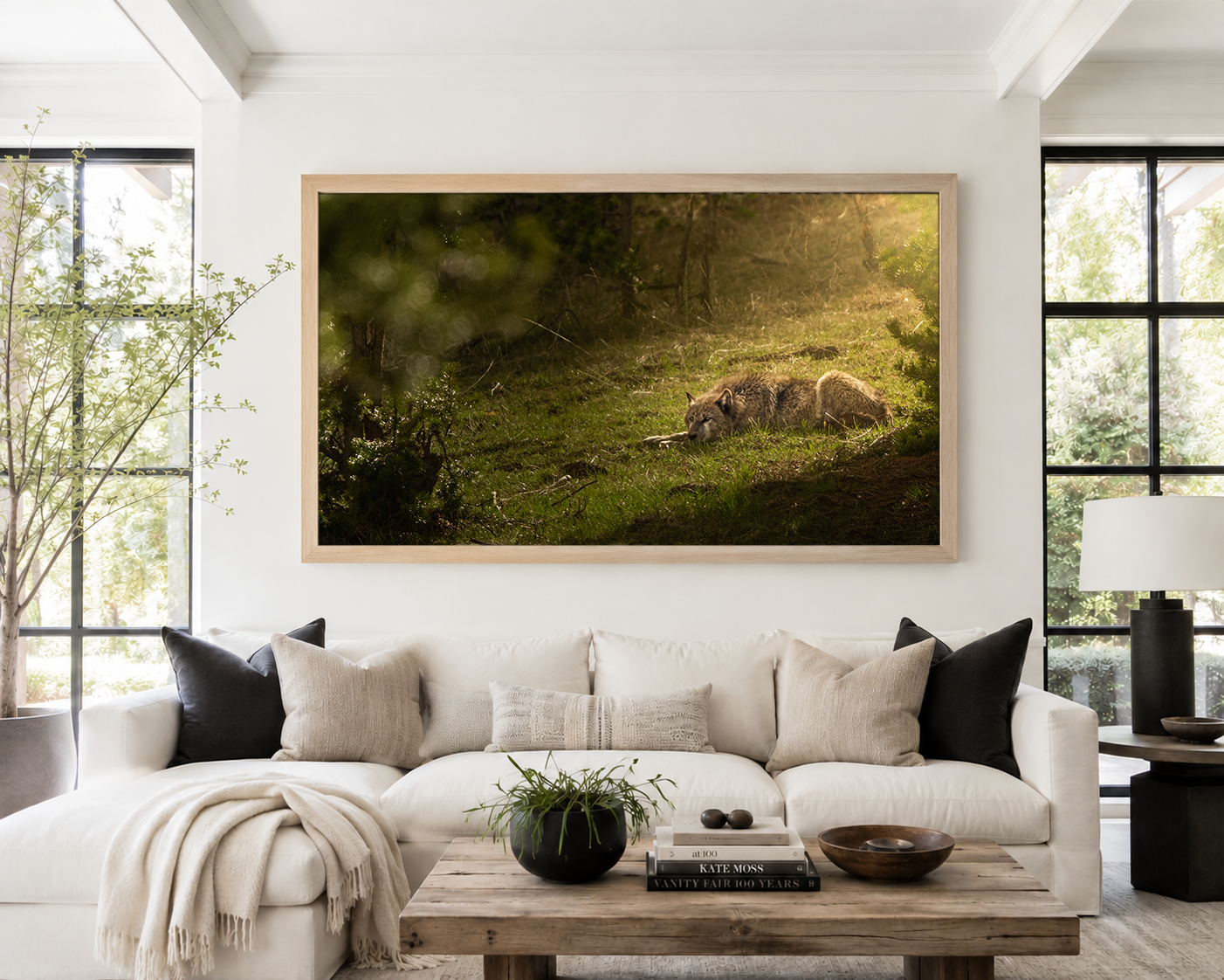 Wide horizontal framed print of a wolf resting in a sunlit forest clearing, hung above a linen sectional in a bright, airy living room.